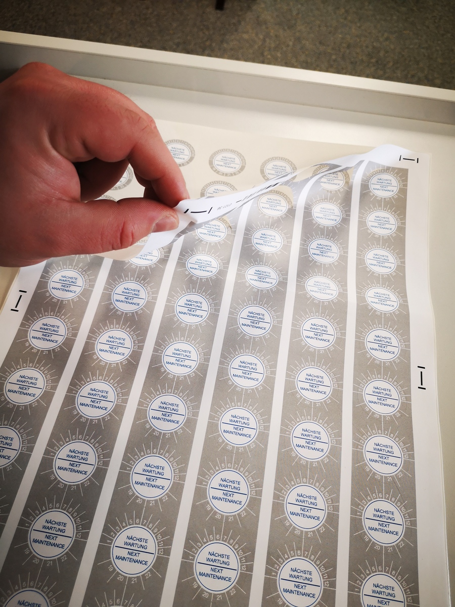 Hand zieht das Schneidegitter von einem Bogen geplotteter runder Prüfplaketten mit Aufschrift Nächste Wartung – Next Maintenance ab