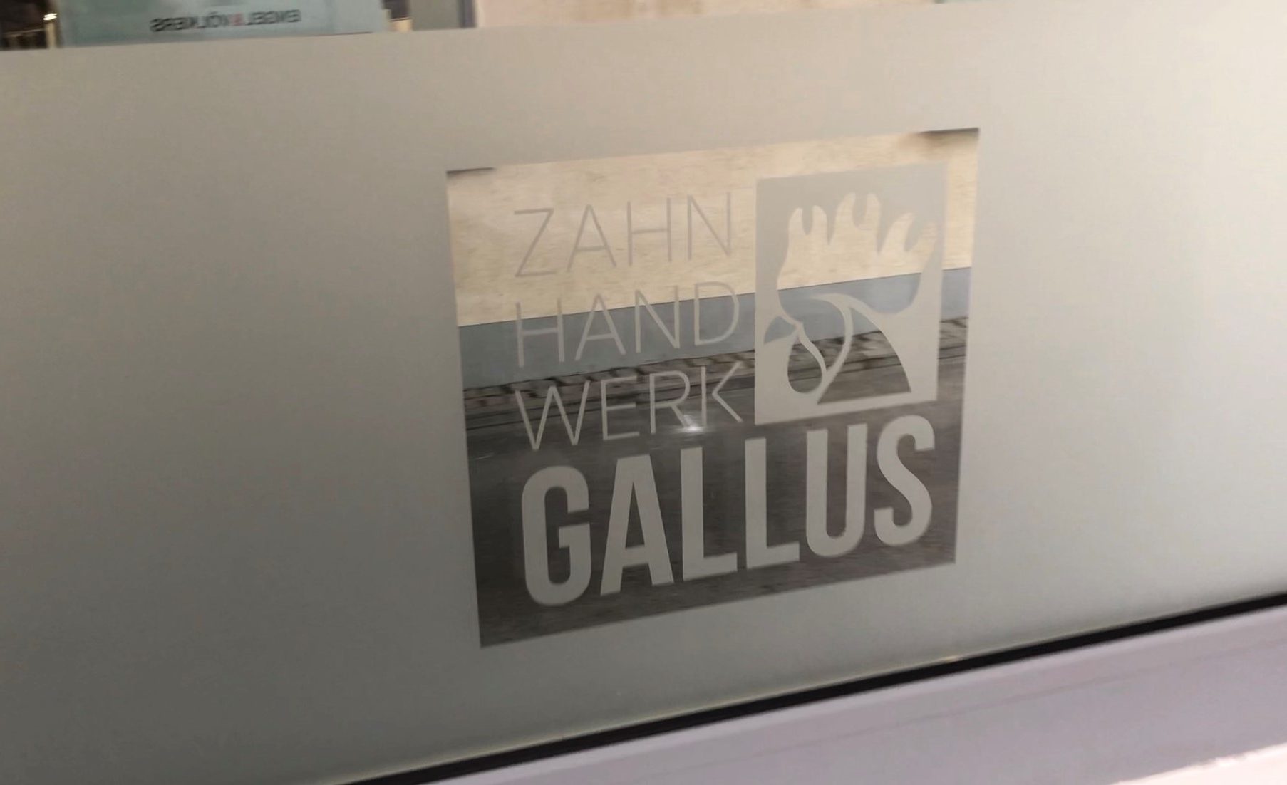 Fenster mit fertig verklebter Milchglasfolie, ausgespartes Logo Zahn Hand Werk Gallus mit Hahnsymbol als klarer Durchblick zur Straße