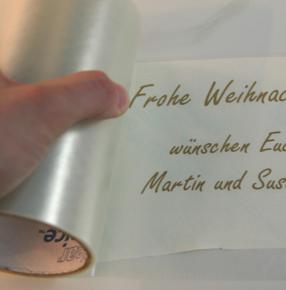 Transparente Applikationsfolie wird von der Rolle auf den geplotteten goldfarbenen Schriftzug Frohe Weihnachten gelegt