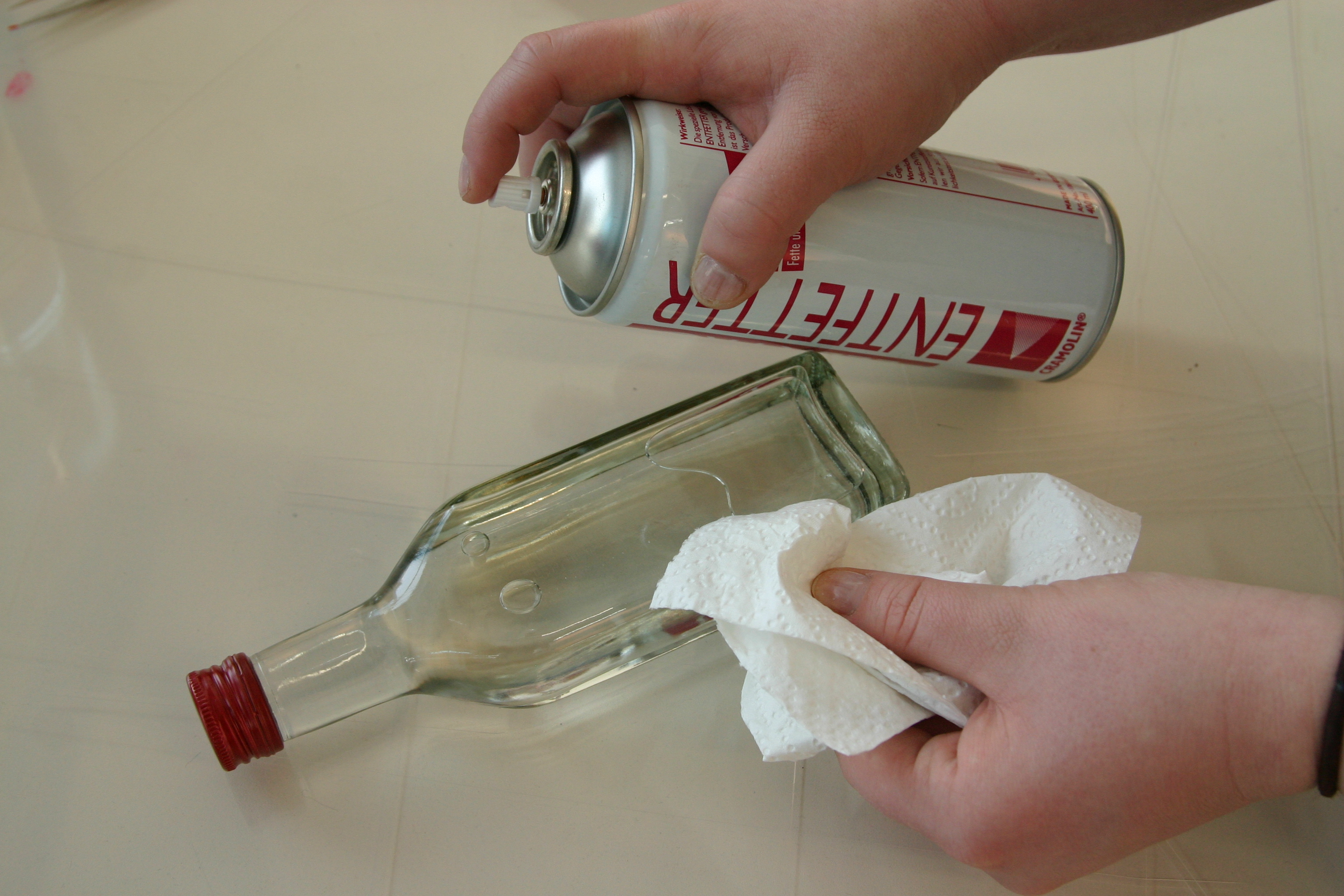 Hand mit Entfetter-Spraydose und Hand mit Küchenpapier reinigen eine leere Glasflasche mit rotem Schraubverschluss, bevor der Schriftzug aufgeklebt wird