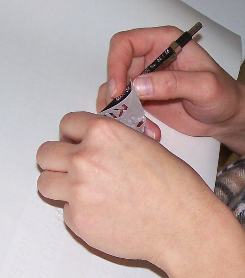 Hände lösen mit einem Druckbleistift überschüssige Milchglasfolie von geplotteten Buchstaben der Bauernregel ab: das Entgittern