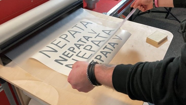 Übertragungsfolie wird vom Spender über entgitterte graue NEPATA-Schriftzüge auf Trägerpapier gezogen, daneben liegt ein Rakel