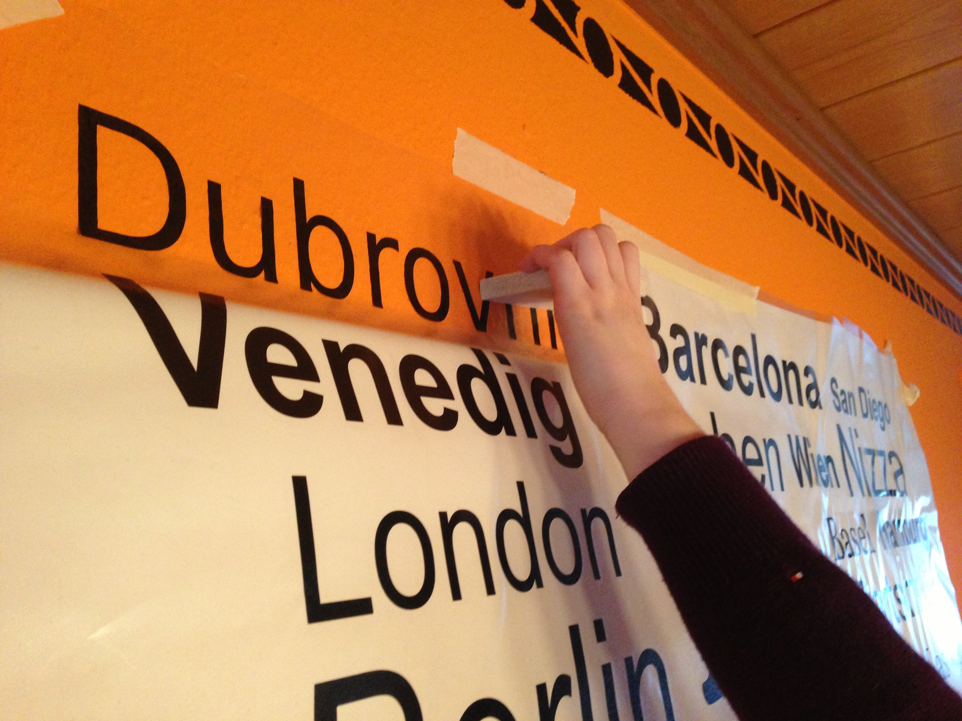Hand rakelt den Schriftzug Dubrovnik mit einer Filzrakel an die orange Wand, darunter hängt noch Silikonpapier mit Städtenamen wie Venedig und London