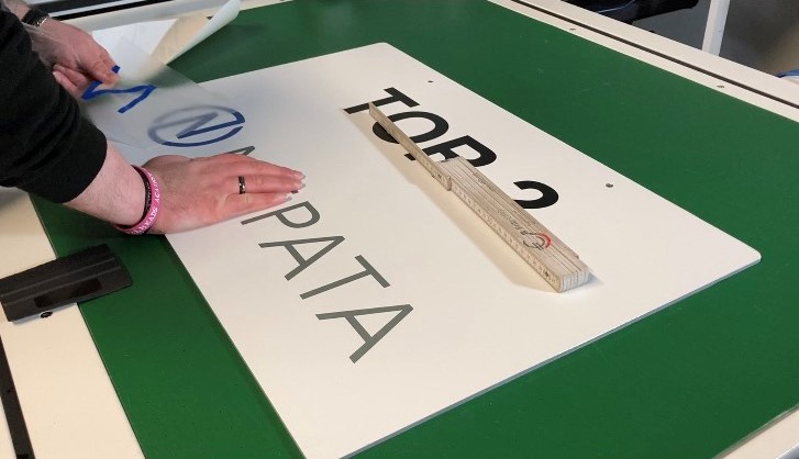 Hände ziehen die Übertragungsfolie von einem weißen Schild mit Aufschrift TOR 2 und Logo ab, darauf ein Zollstock, daneben ein Rakel
