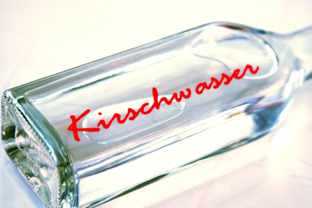 Liegende Glasflasche mit roter Schreibschrift Kirschwasser aus Flockfolie, die auch über die geprägten Vertiefungen im Glas verläuft
