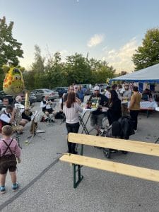 Gäste und Kind in Lederhose bei der Nepata Wiesn 2021 im Freien, Blasmusiker spielen neben Bierbank und Zelt in der Abendsonne