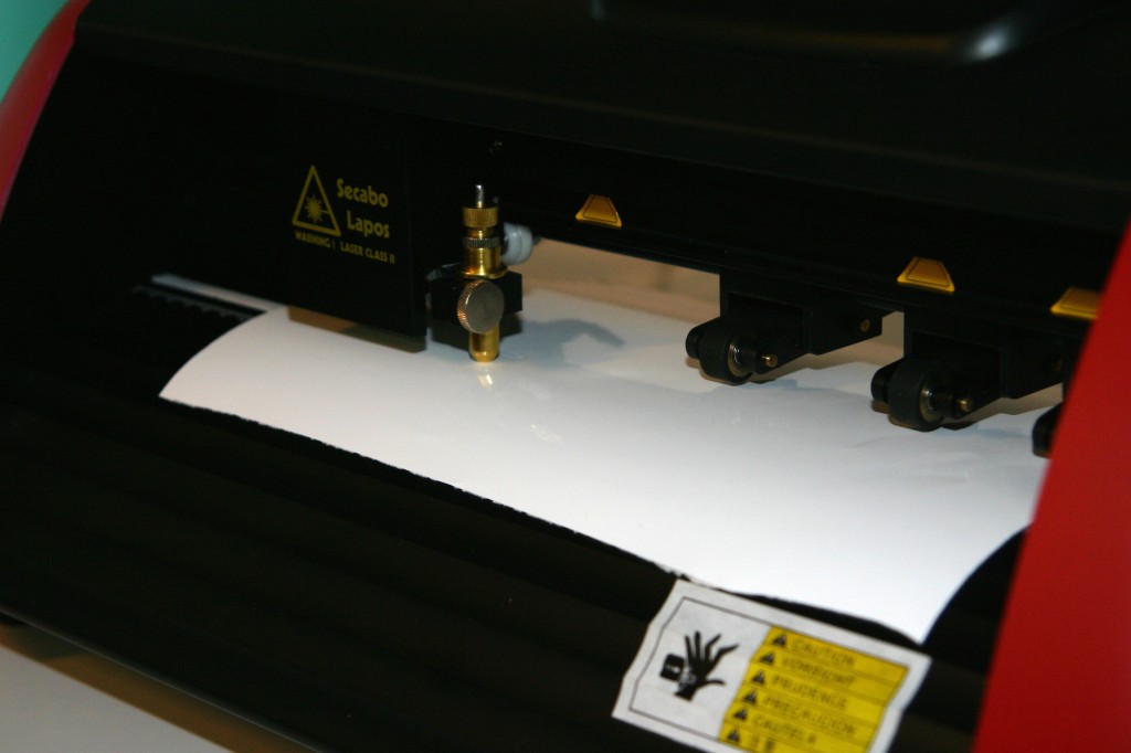 Schneideplotter Secabo Lapos mit Messerhalter und Andruckrollen über weißer Selbstklebefolie, bereit zum Plotten des Schriftzugs