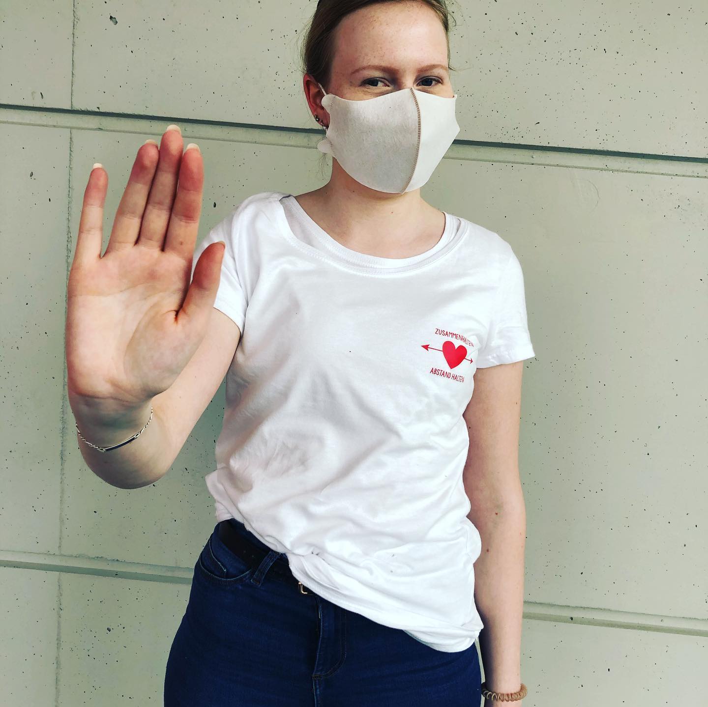 Frau mit Maske zeigt Stopp-Geste, trägt weißes T-Shirt mit rotem Herz-Pfeil-Motiv und Aufdruck Zusammenhalten Abstand halten