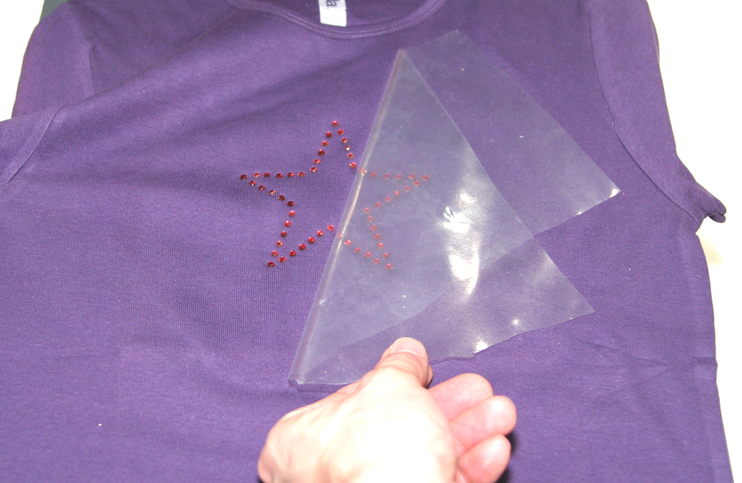 Hand zieht transparente Übertragungsfolie von einem Stern aus roten Strass-Steinen auf einem lila T-Shirt ab – letzter Arbeitsschritt