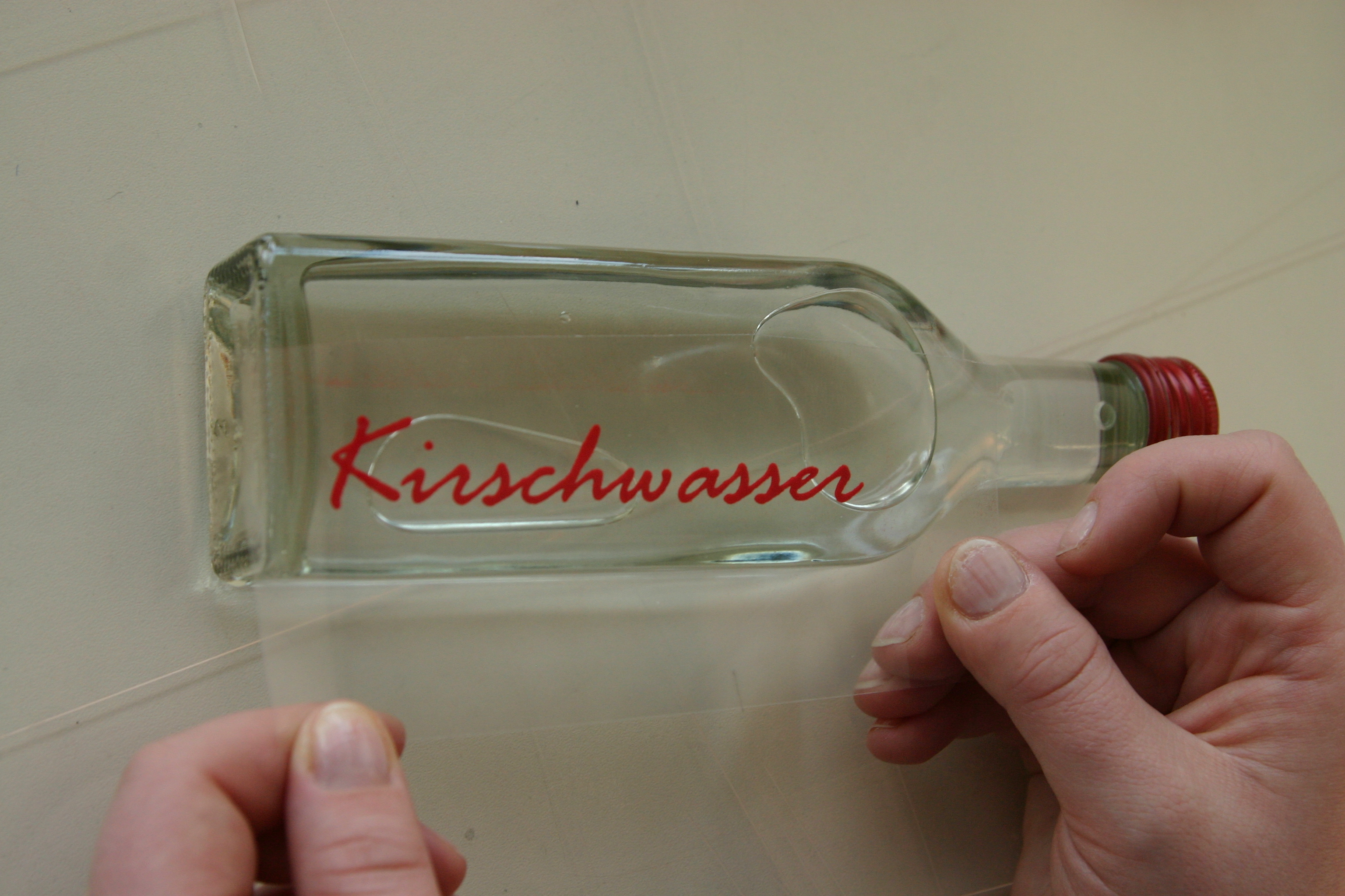 Hände ziehen die transparente Übertragungsfolie von einer Glasflasche mit Schraubverschluss ab, darauf klebt der rote Schriftzug Kirschwasser