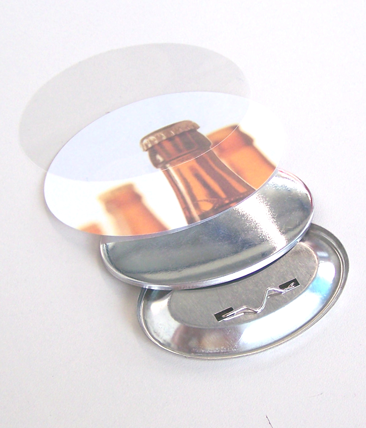 Einzelteile eines ovalen Buttons: Klarsichtfolie, Motivpapier mit Bierflaschen, Vorderschale aus Metall und Rückwand mit Anstecknadel