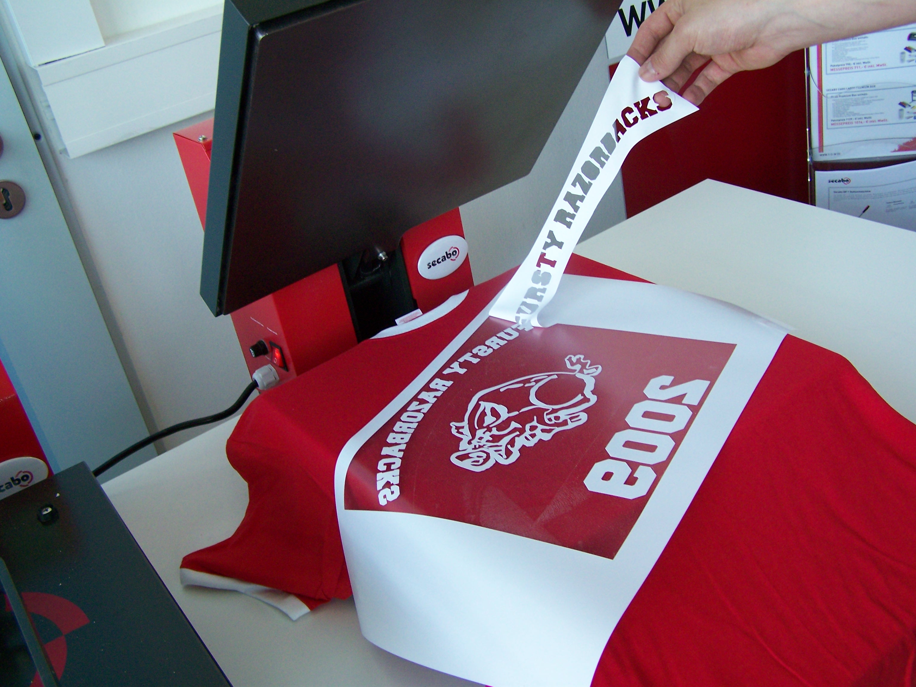 Hand zieht nach dem Pressen die Trägerfolie vom roten T-Shirt ab, darunter weißes Folienmotiv Varsity Razorbacks 2009, Transferpresse von secabo