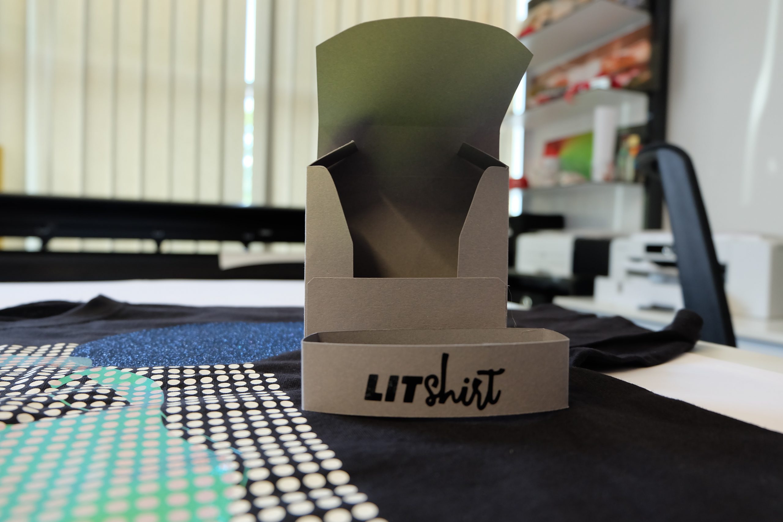 Gefaltete Verpackung aus grauem Karton mit schwarzem LITshirt-Logo aus Folie, auf einem schwarzen T-Shirt mit Punktemuster