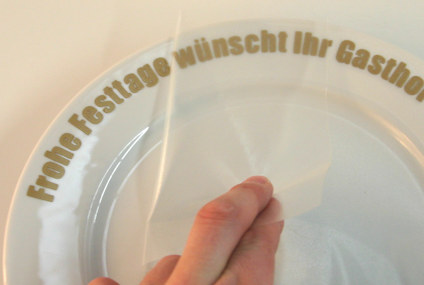 Hand zieht Übertragungsfolie von weißem Teller ab, am Rand der goldene Folienschriftzug Frohe Festtage wünscht Ihr Gasthof…