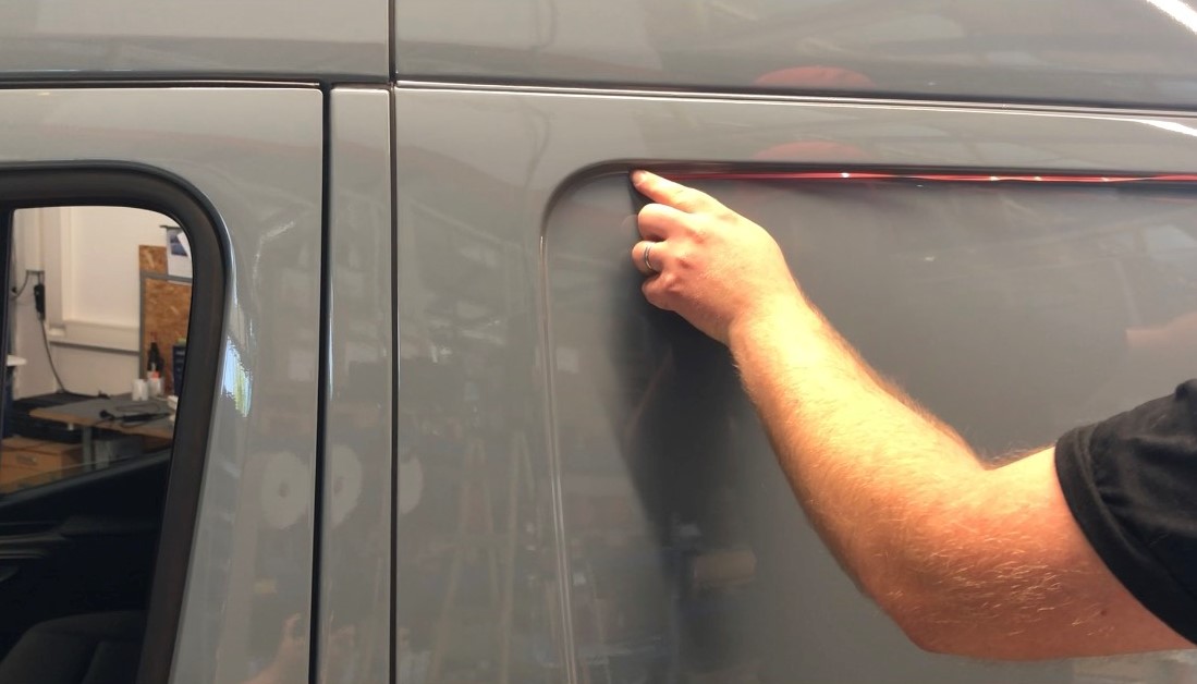 Hand zieht rotes Schneidetape entlang einer Kante an der grauen Seitenwand eines Transporters, um die Folie ohne Cuttermesser zu durchtrennen