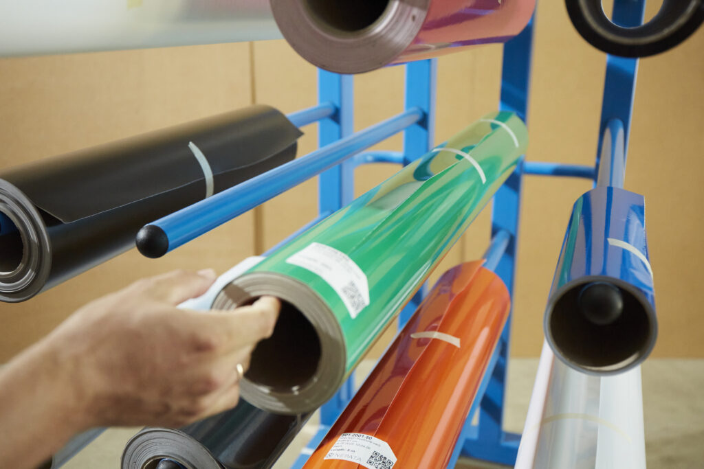 Hand zieht grüne Vinylrolle von einer Stange des blauen Nepata-Rollentransportwagens, daneben schwarze, orange und blaue Folienrollen