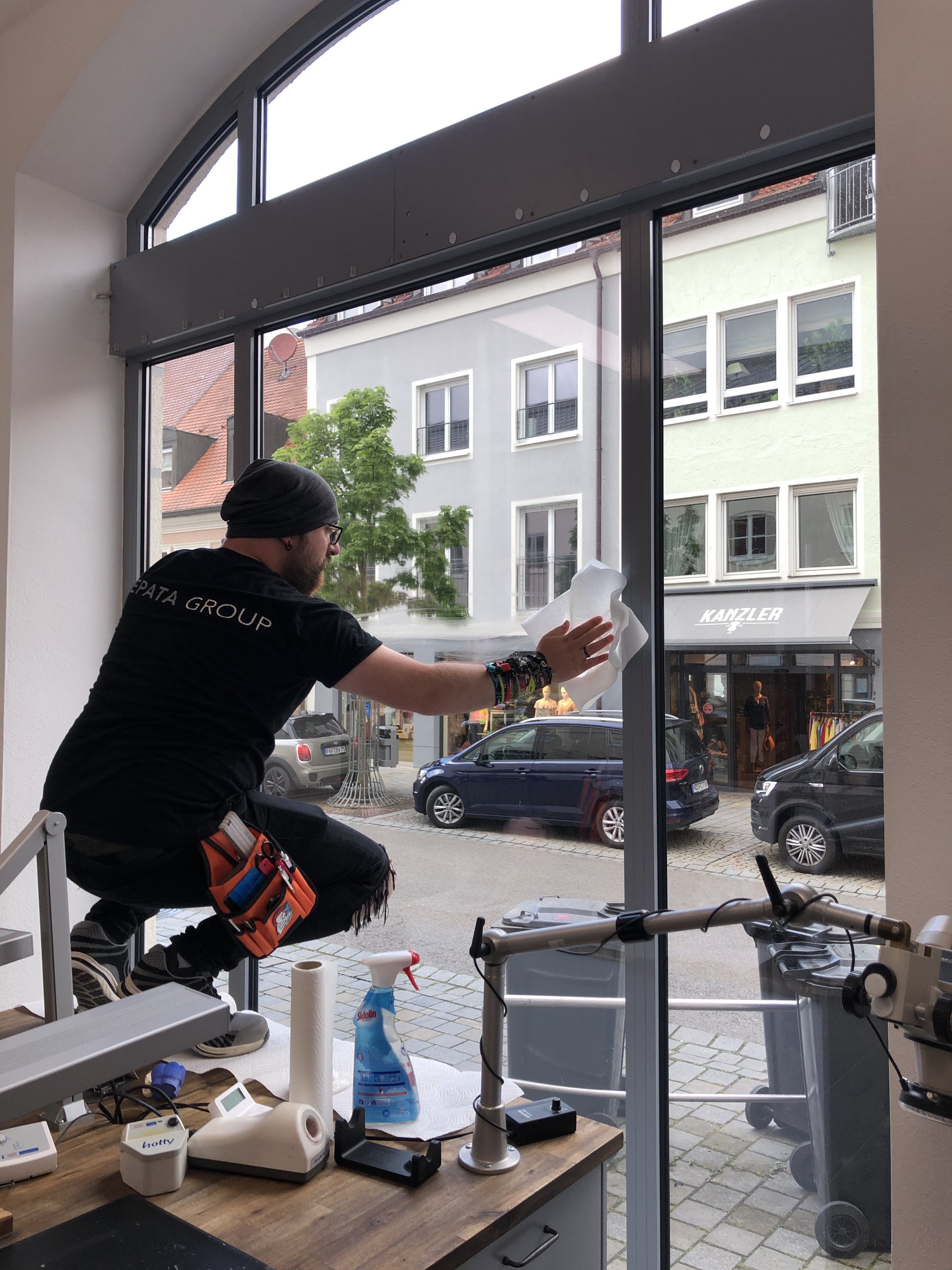 Mann mit Werkzeuggürtel reinigt große Schaufensterscheibe mit Küchenpapier vor dem Folieren, daneben Glasreiniger-Sprühflasche und Küchenrolle