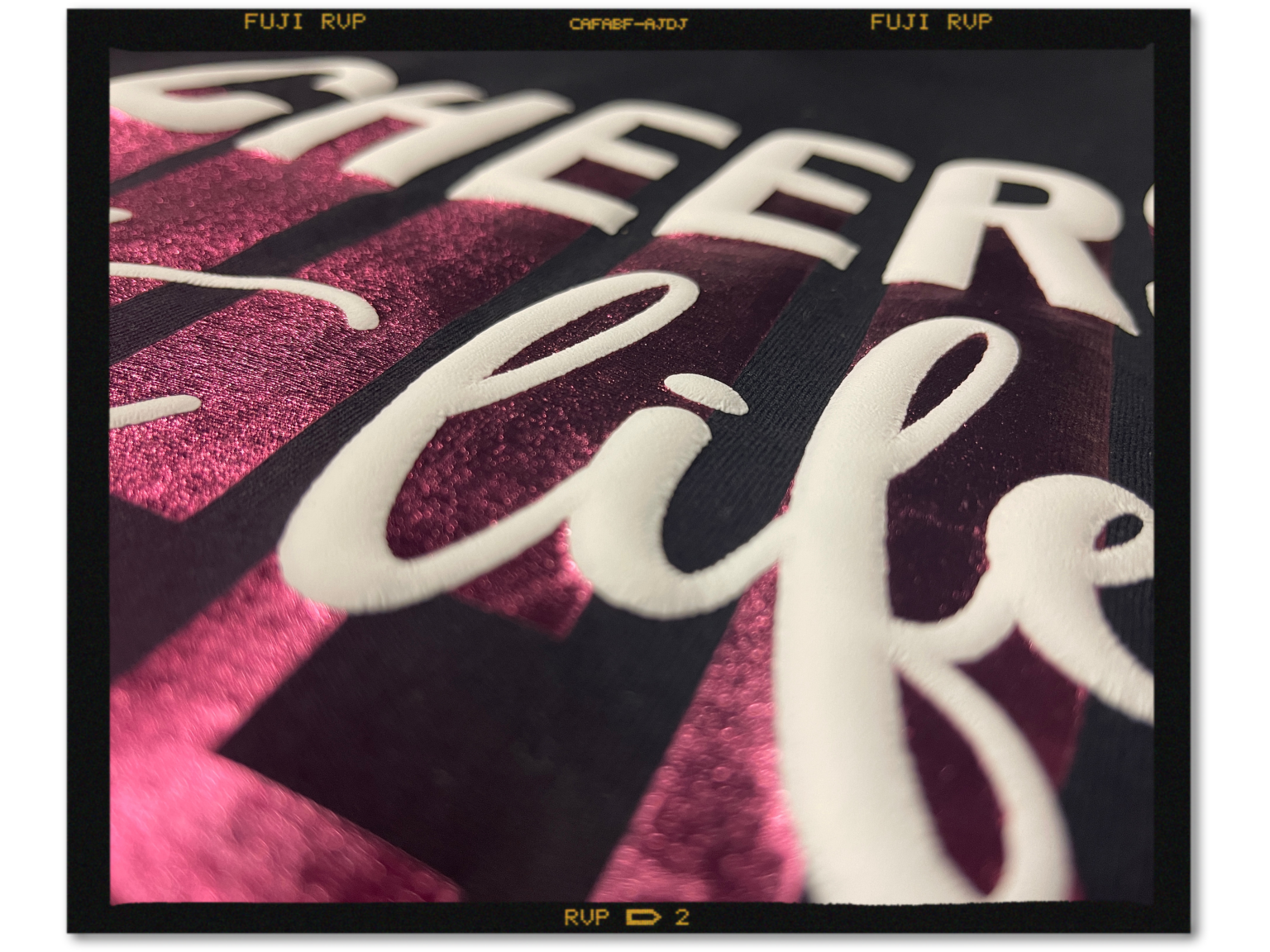Nahaufnahme eines schwarzen T-Shirts: pinke Metallic-Flexfolie mit weißem 3D-Puff-Schriftzug Cheers to life, erhabene Buchstaben