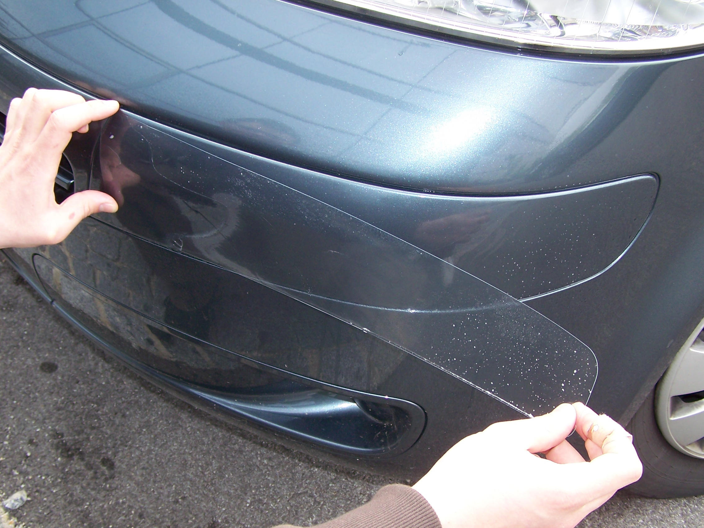 Zwei Hände legen eine zugeschnittene, transparente Steinschlagschutzfolie auf die nass eingesprühte Stoßstange eines dunkelgrauen Autos