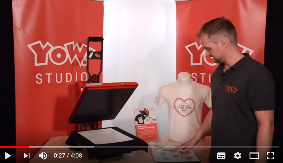 Videostandbild YOW! Studio: Mitarbeiter zeigt Transferbogen mit Motiven neben geöffneter Transferpresse und bedrucktem T-Shirt
