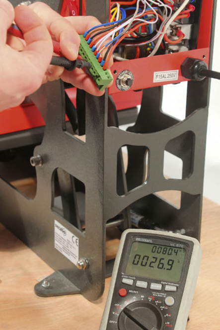 Hände prüfen mit Multimeter die Klemmleiste eines geöffneten roten Geräts, am schwarzen Gestell ein CE-Typenschild: elektrische Qualitätsprüfung