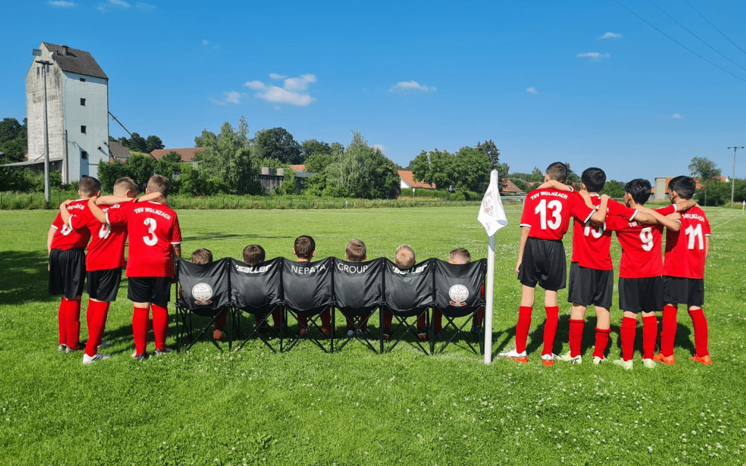 TSV Wolnzach’s D2 team in red, seen from behind; children on a black folding bench bearing the TSV logo and ‘Nepata Group’ lettering at the edge of the pitch