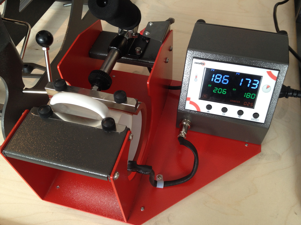 Secabo TM1 Tassenpresse mit eingespannter weißer Tasse, Display zeigt 186 °C bei Soll 206 °C und 173 von 180 Sekunden Pressdauer