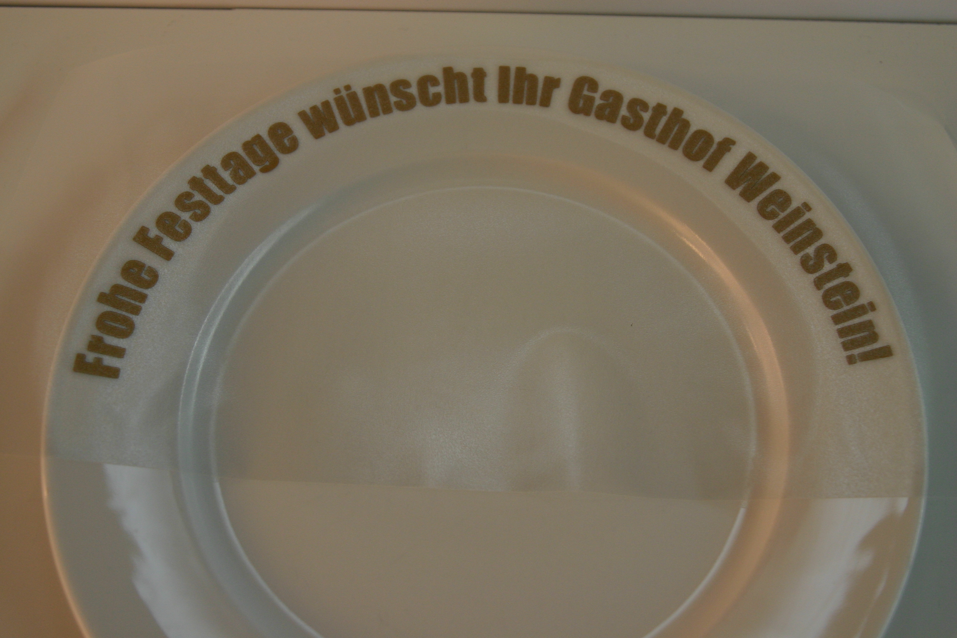 Weißer Teller mit goldenem Folienschriftzug Frohe Festtage wünscht Ihr Gasthof Weinstein! am Rand, noch mit Übertragungsfolie überzogen