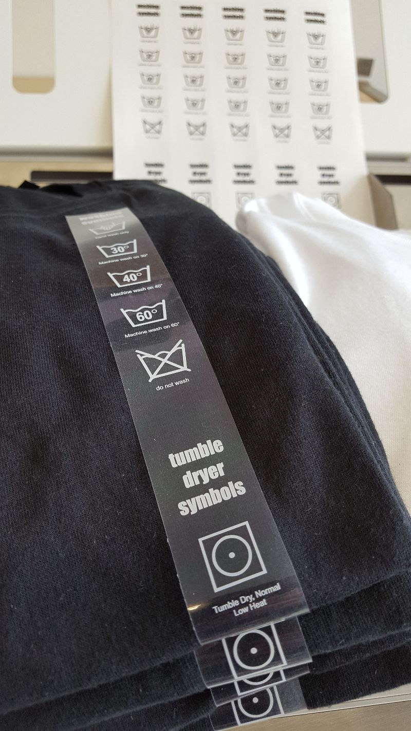 Transparenter Textilaufkleber mit Wasch- und Trocknersymbolen auf schwarzem Shirt, dahinter ein Bogen weiterer Aufkleber mit Pflegesymbolen