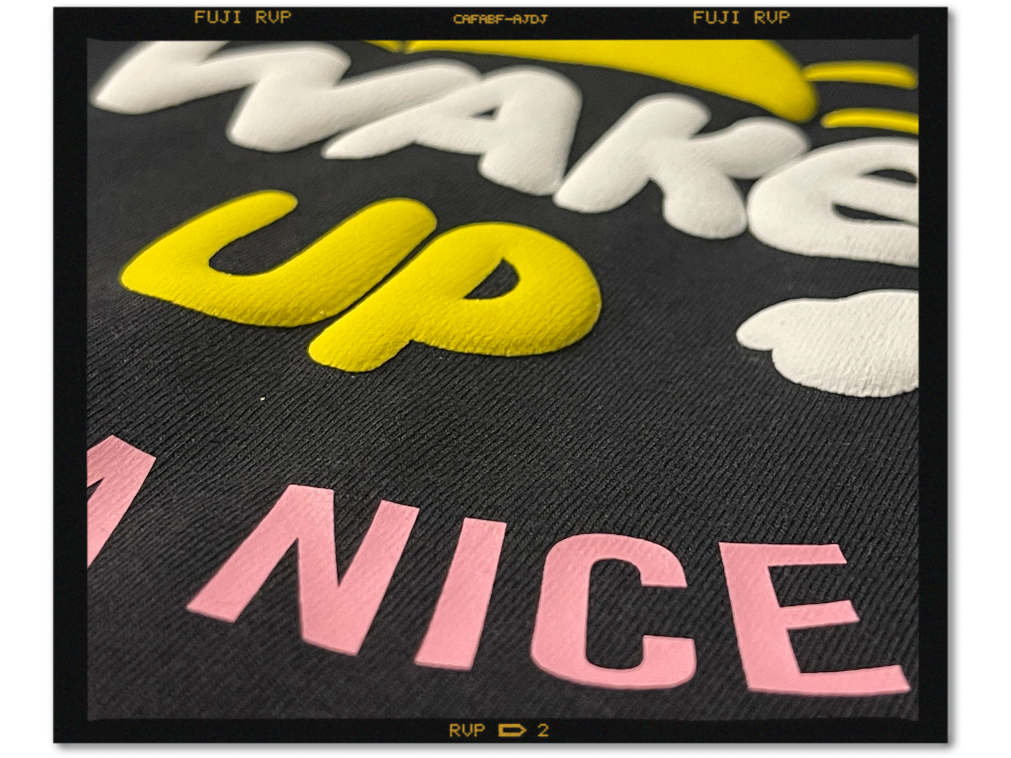 Nahaufnahme eines schwarzen Shirts: Wake Up erhaben aus Puff-Folie in Weiß und Gelb, darunter Nice flach in FlexCut Candy Pink