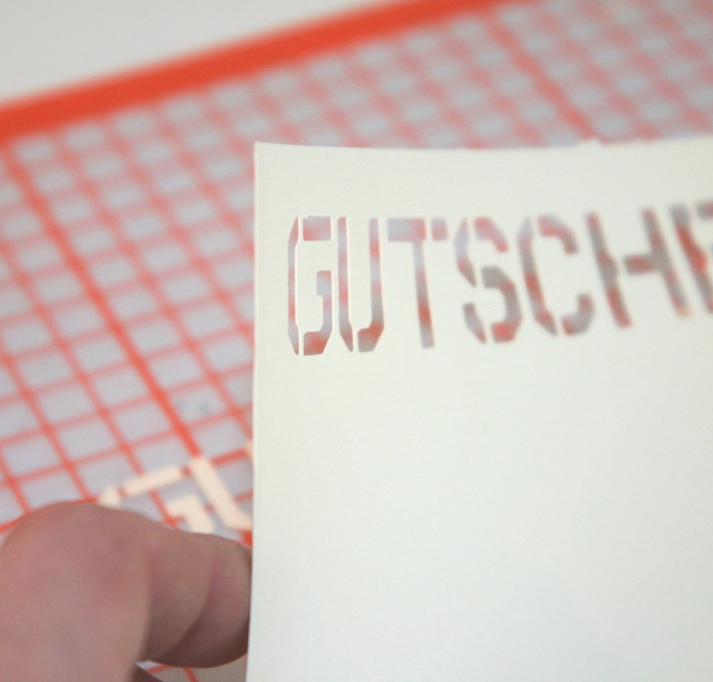 Hand zieht hellen Karton mit ausgeplottetem Schriftzug Gutschein in Schablonenschrift von orangefarbener Plotunterlage ab