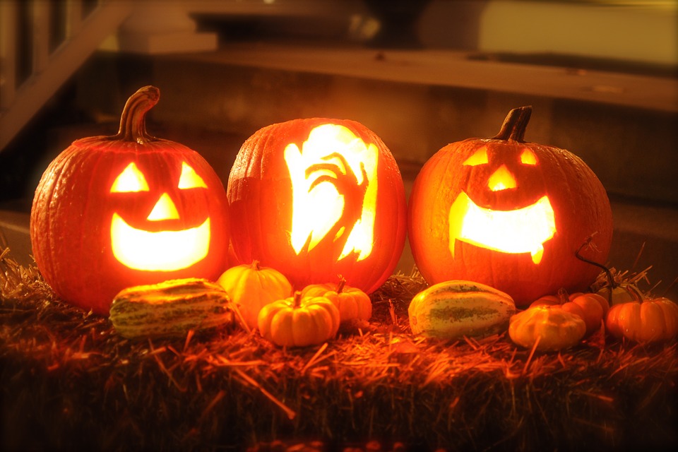 Drei leuchtende geschnitzte Halloween-Kürbisse mit Gesichtern und Hand-Motiv, davor kleine Zierkürbisse auf einem Strohballen