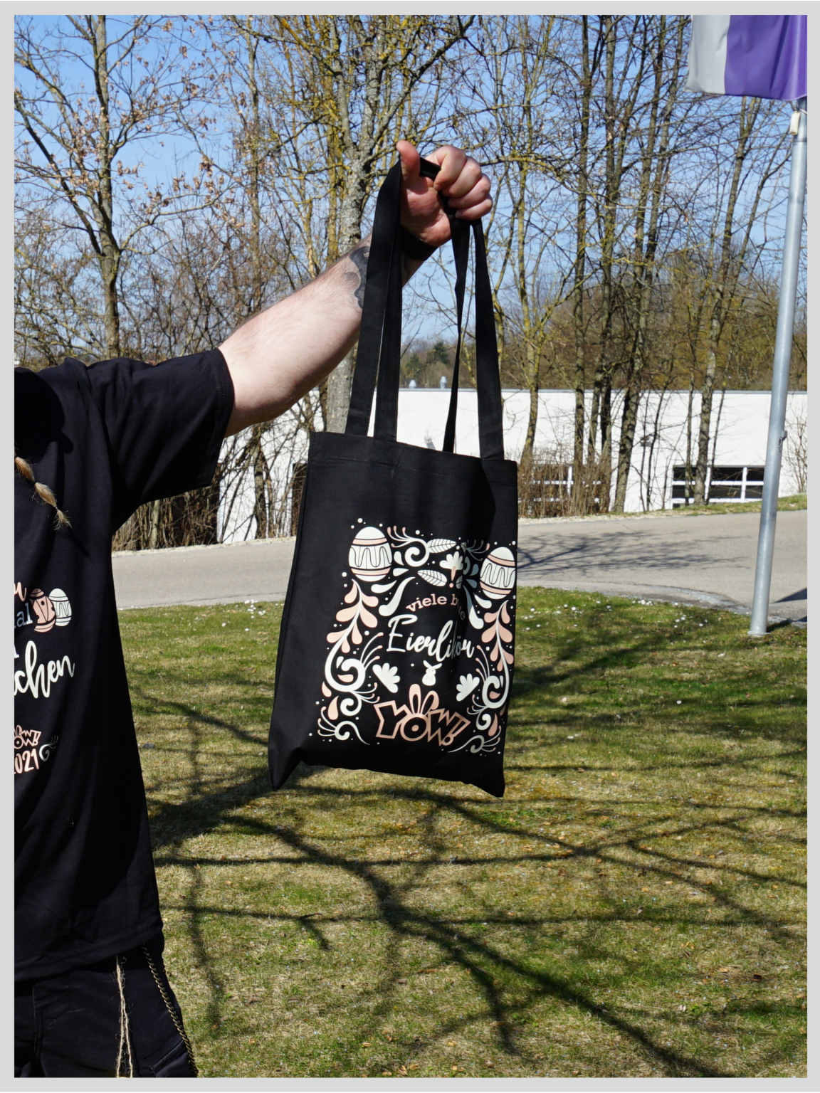 Mit Flexfolie veredelte schwarze Stofftasche mit Ostermotiv aus Eiern, Ornamenten und YOW!-Logo in Weiß und Rosé, daneben ein passendes Shirt