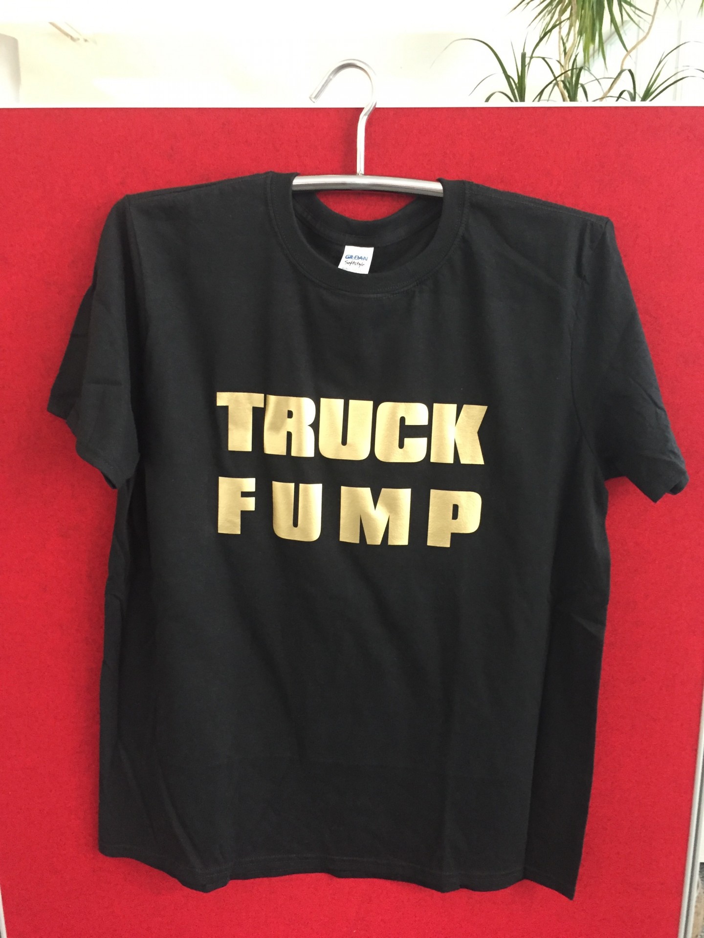 Fertiges schwarzes T-Shirt mit Schriftzug TRUCK FUMP aus goldener Flexfolie, auf einem Kleiderbügel vor roter Stellwand
