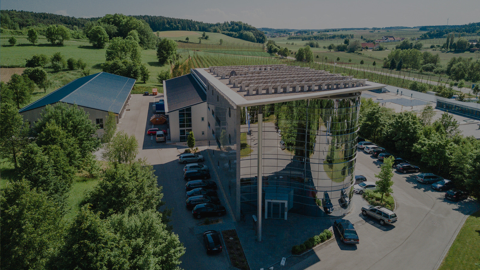 Vue aérienne d'un bâtiment d'entreprise doté d'une façade vitrée arrondie, de places de parking et d'entrepôts, niché au milieu de collines verdoyantes et de champs de houblon