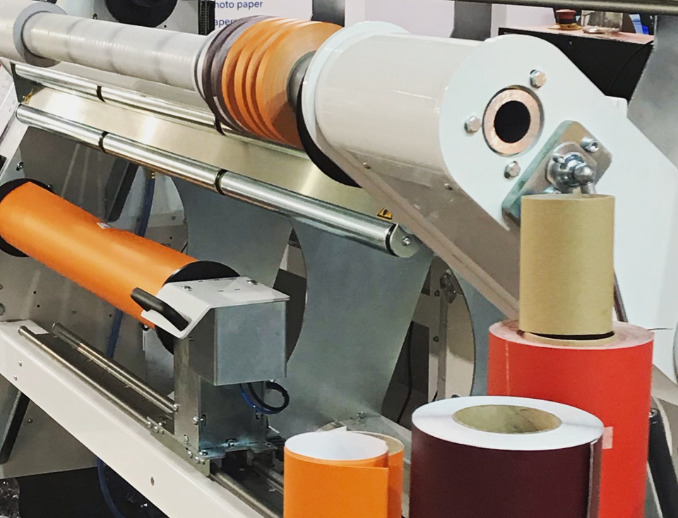 Umwickelmaschine mit orangefarbener Folienrolle, davor rote, weinrote und orange Folienrollen und Pappkerne griffbereit abgestellt