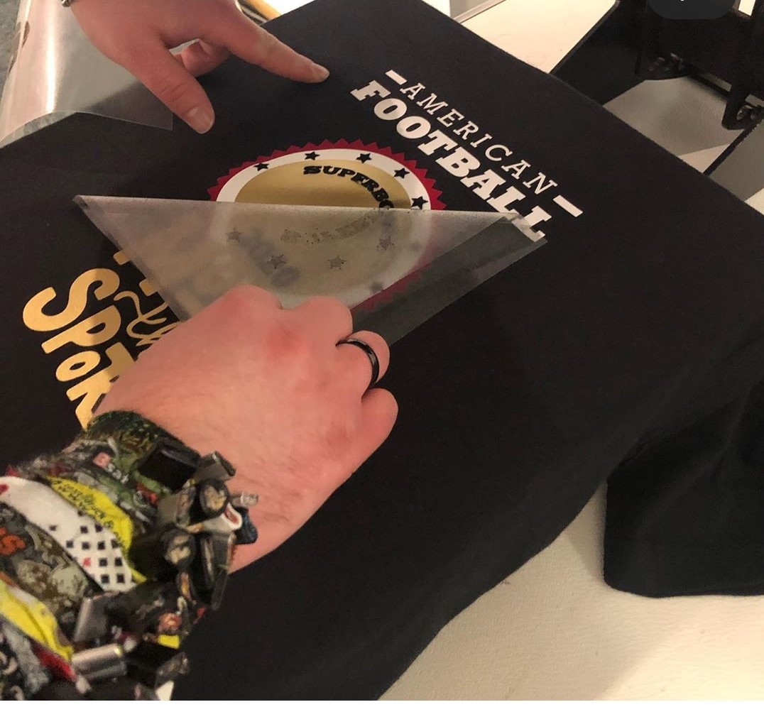 Hände ziehen die Trägerfolie von einem schwarzen Shirt mit frisch aufgepresstem American-Football-Superbowl-Motiv in Gold, Weiß und Rot ab