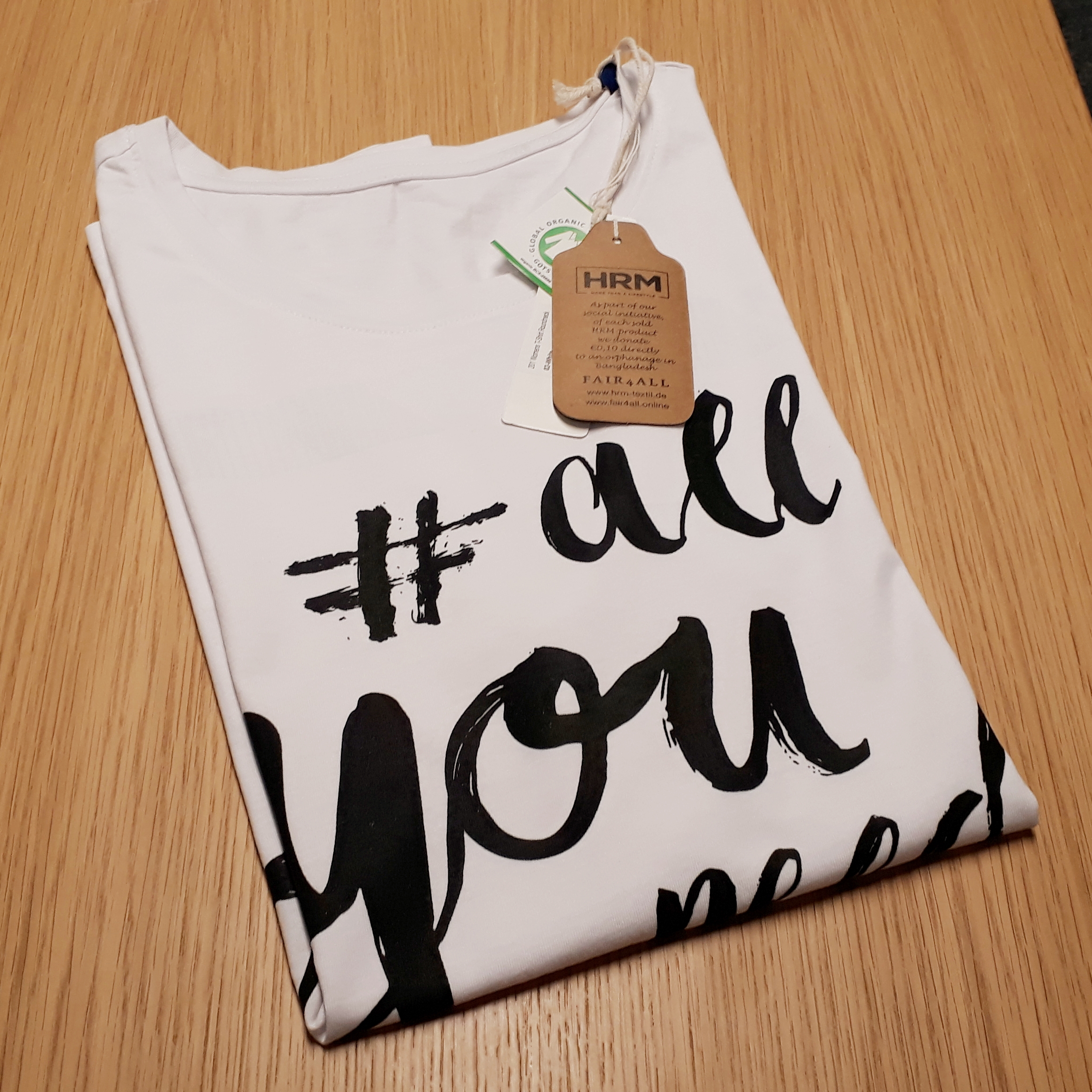 Gefaltetes weißes T-Shirt, am Kragen per Schnur befestigtes HRM-Anhängeetikett aus Kraftpapier und GOTS-Etikett, kleberlos auf Textil