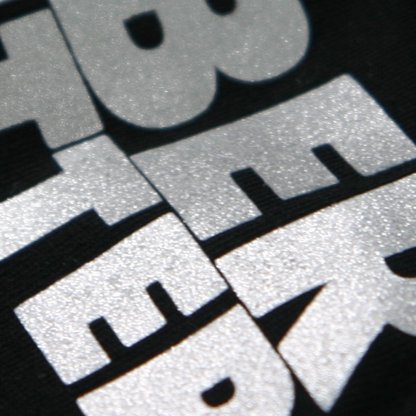 Nahaufnahme silbergrauer Flexfolie mit feinkörnig glänzender Oberfläche, als Blockbuchstaben auf schwarzes Textil gepresst