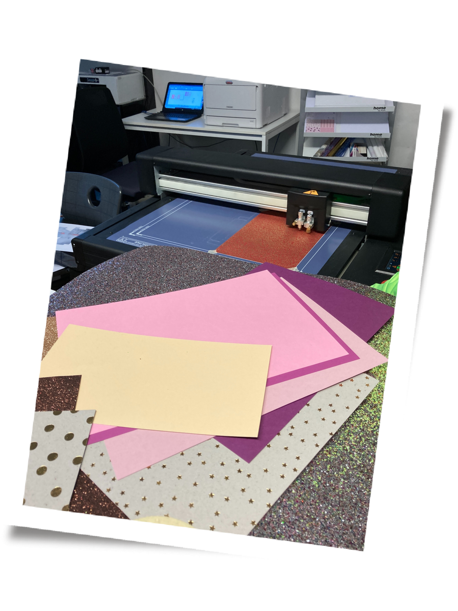 Schneideplotter Vulcan FC-500VC mit rotem Glitzerpapier, davor bunte Glitzer- und Designpapiere mit Goldsternen; im Hintergrund Laptop mit DrawCut