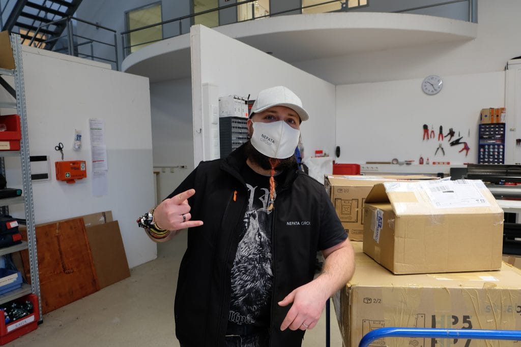 Anwendungstechniker Ääd mit Nepata-Maske und weißer Kappe zeigt die Rock-Geste in der Werkstatt neben gestapelten Paketen