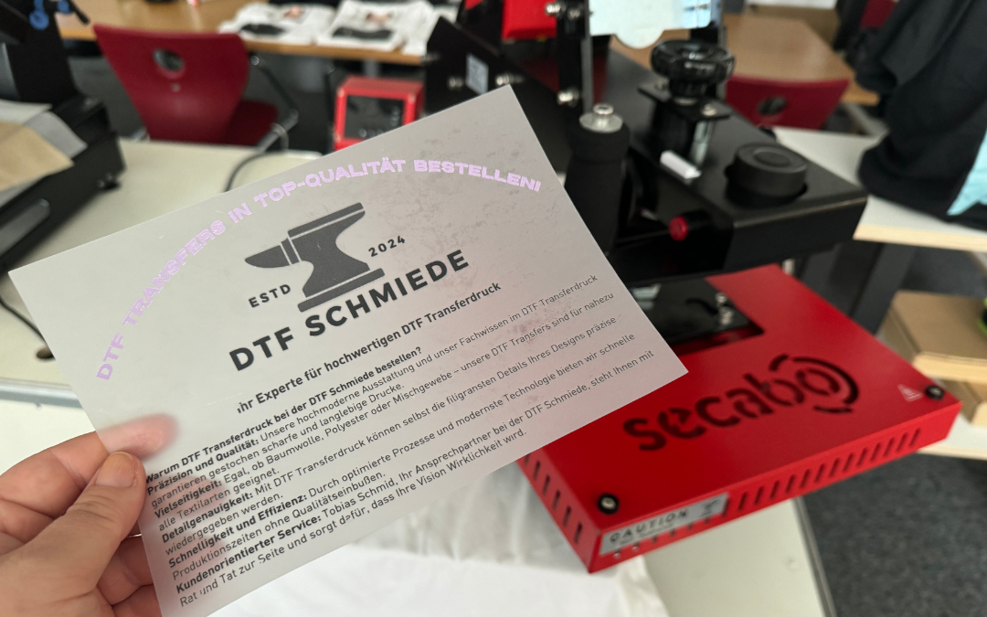 Hand hält DTF-Transfer der DTF-Schmiede mit Amboss-Logo und Werbetext vor einer roten Secabo-Transferpresse