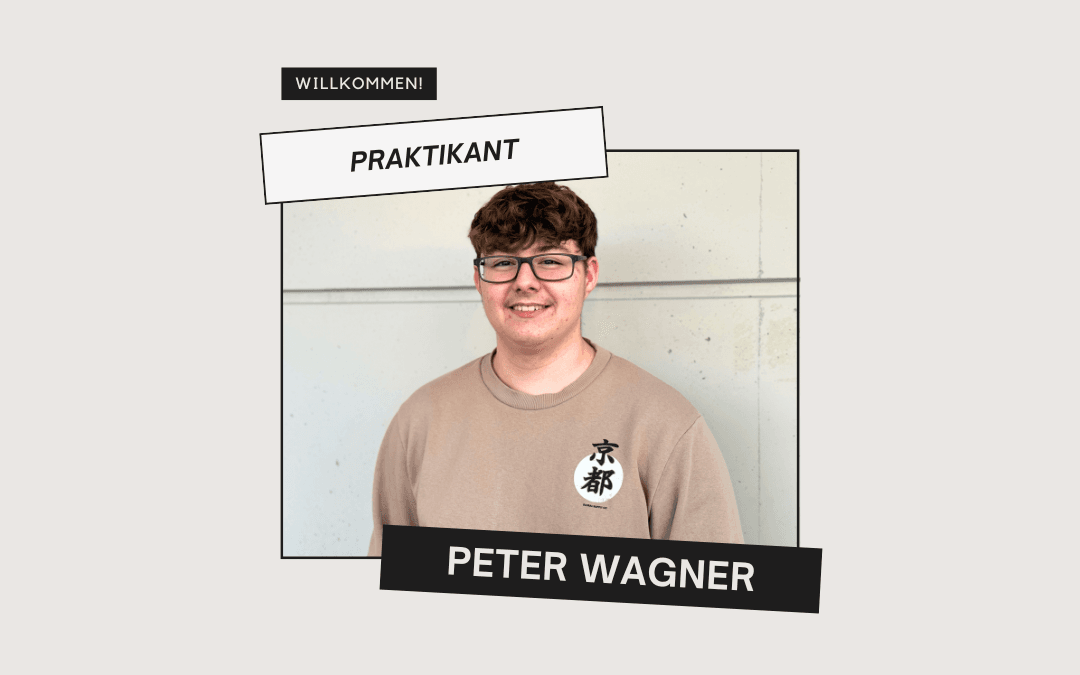 Welcome image: Intern Peter Wagner, wearing glasses and a beige jumper, smiles in front of a light-coloured concrete wall bearing the word ‘Intern’ and his name