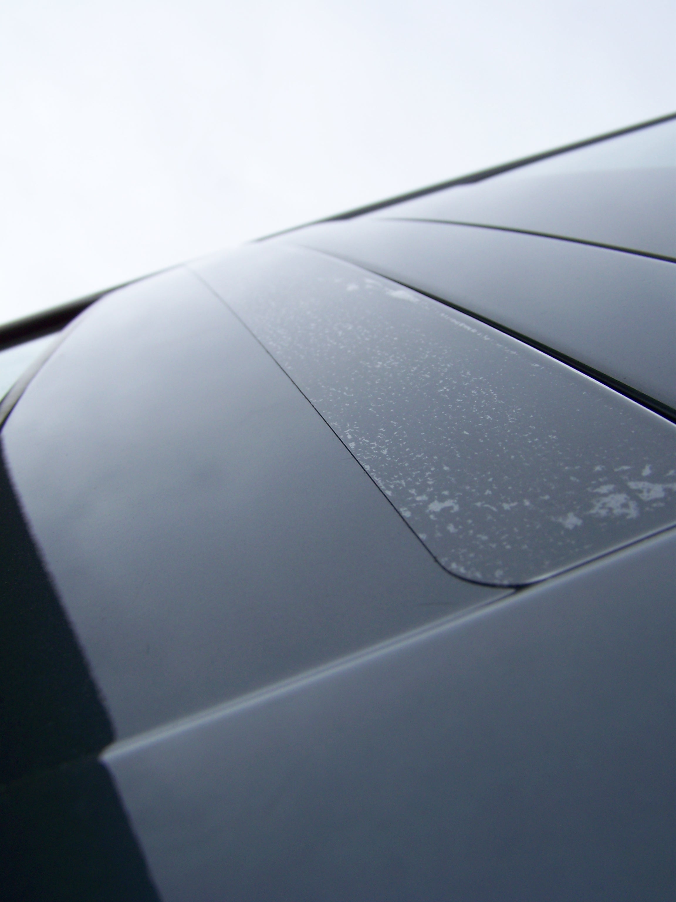 Nahaufnahme einer dunklen Autokarosserie mit aufgeklebtem, abgerundetem Streifen transparenter Steinschlagschutzfolie, darauf helle Flecken