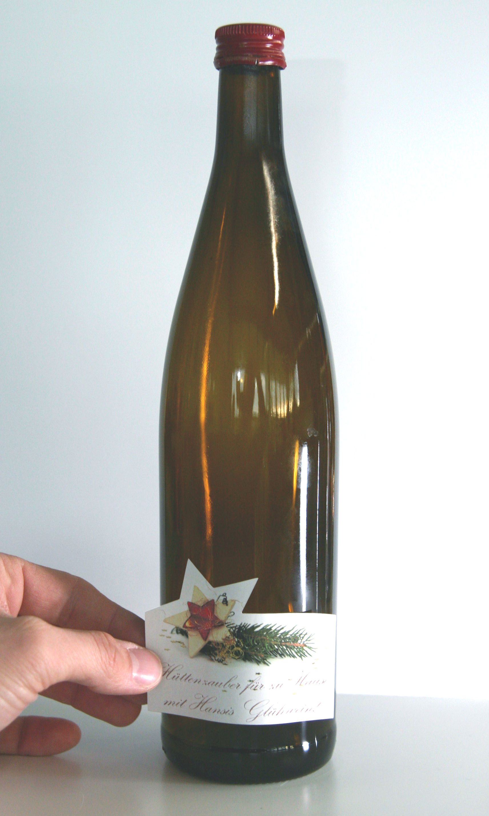 Hand bringt geplottetes Etikett mit Stern, Tannenzweig und Aufschrift Hüttenzauber für zu Haus mit Hansis Glühwein auf braune Flasche auf