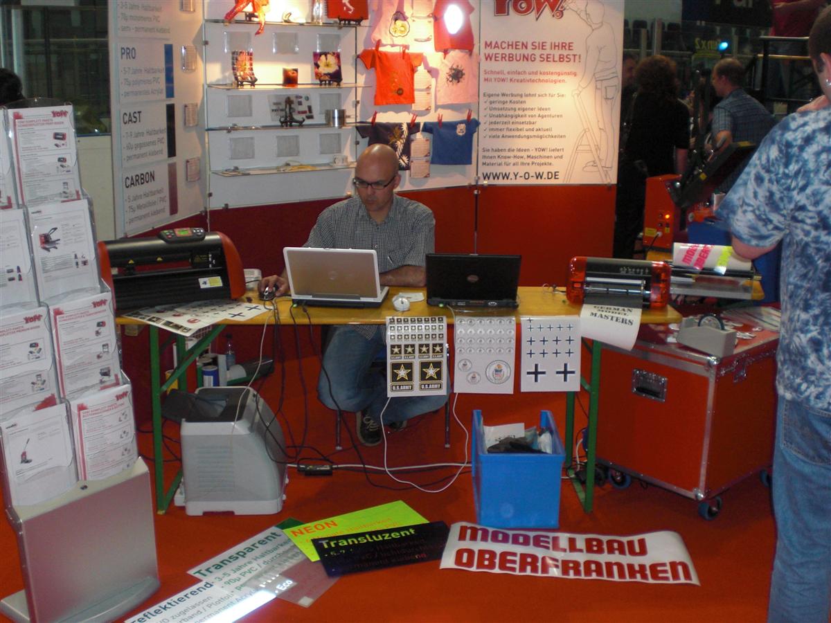 YOW!-Messestand auf der German Model Masters in Nürnberg: Mitarbeiter am Laptop mit Schneideplottern, Modellbau-Aufklebern und Folienmustern