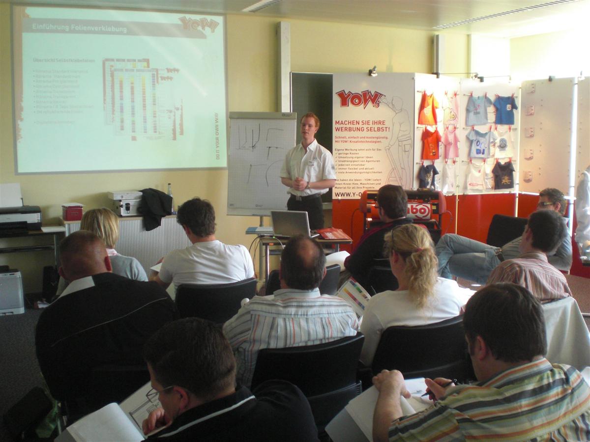 YOW!-Workshop zur Folienverklebung: Referent vor Flipchart und Präsentation, Teilnehmende an Tischen, daneben Werbewand mit bedruckten T-Shirts