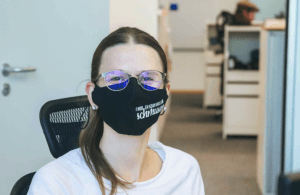 Mitarbeiterin mit Brille und schwarzer Mund-Nasen-Maske mit weißem Aufdruck sitzt an ihrem Arbeitsplatz im Büro