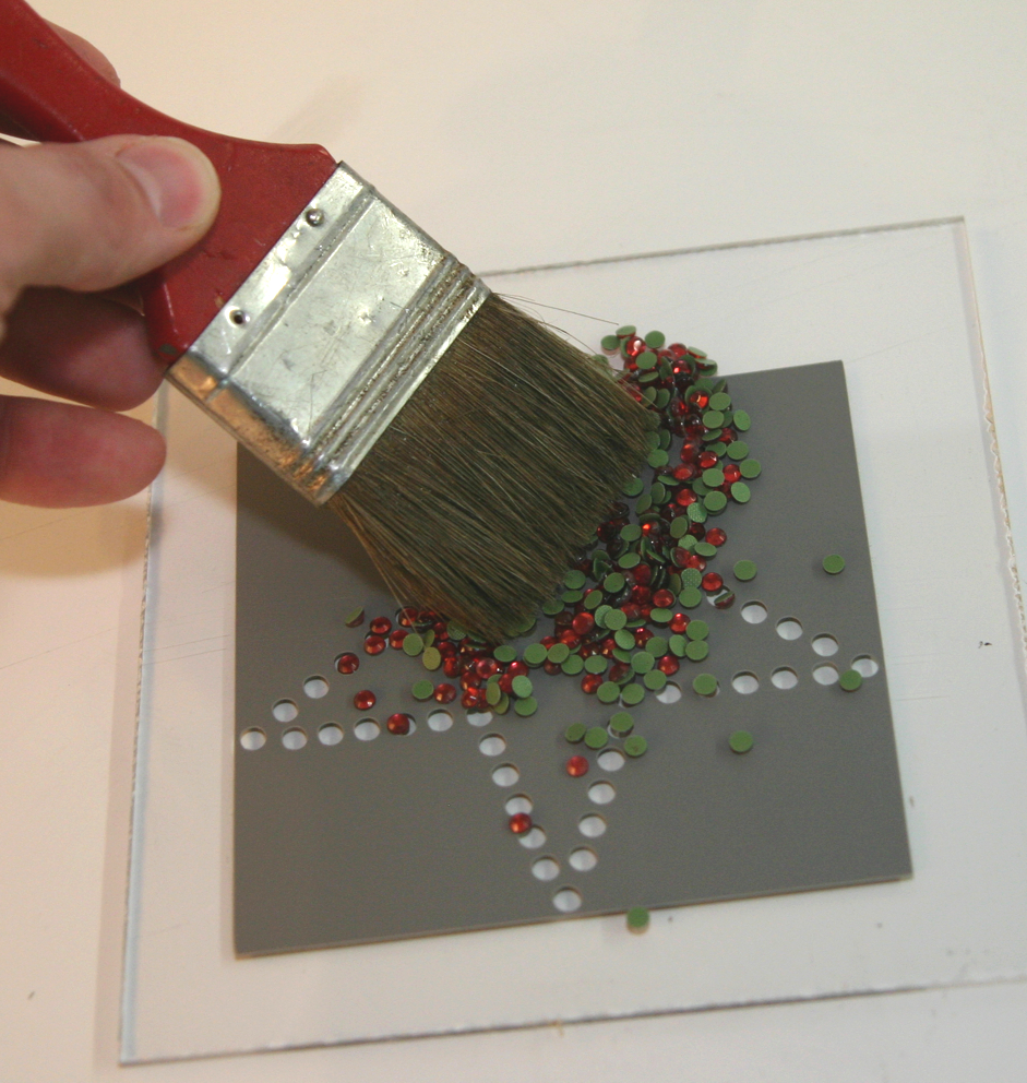 Pinsel wischt rote Strass-Steine über eine graue geplottete Schablone auf Acrylglas, damit sie in die Löcher des Motivs fallen
