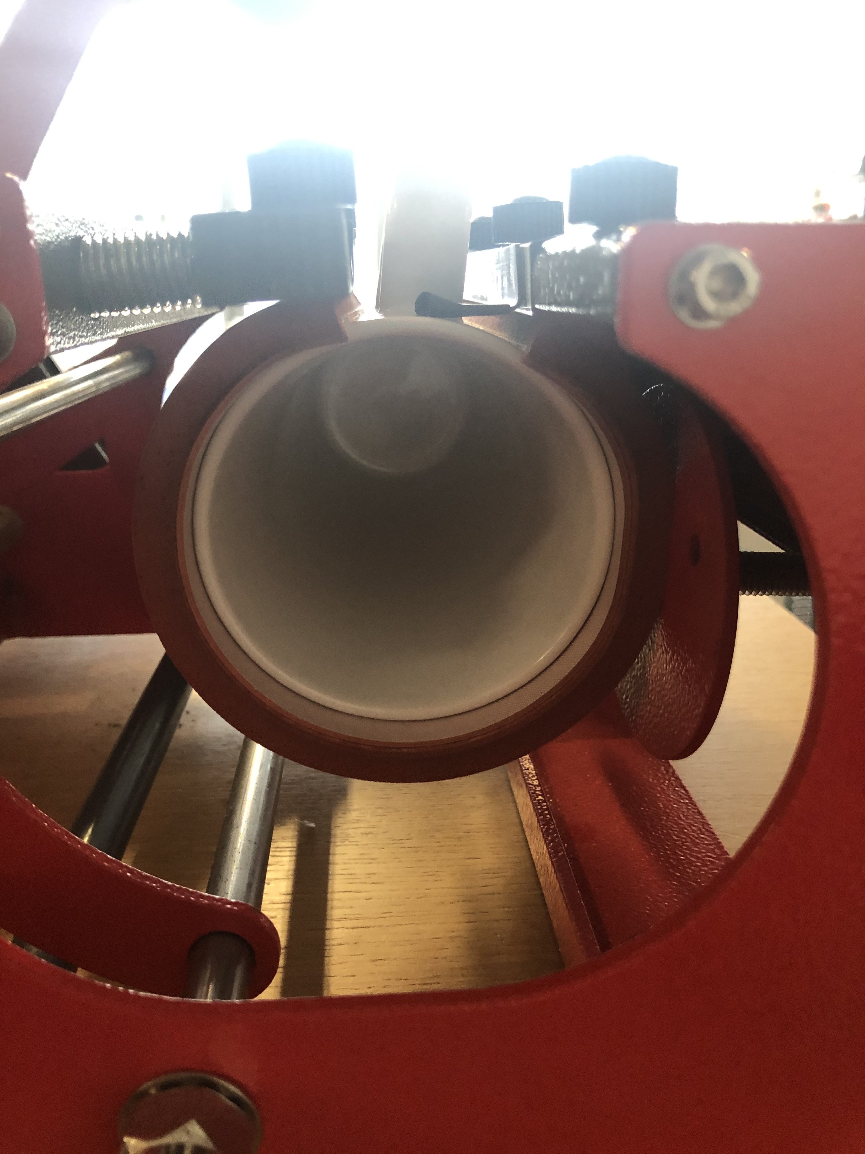 Blick in eine weiße Tasse, eingespannt in der Heizmanschette einer roten Tassenpresse, dahinter die schwarzen Stellschrauben für den Anpressdruck